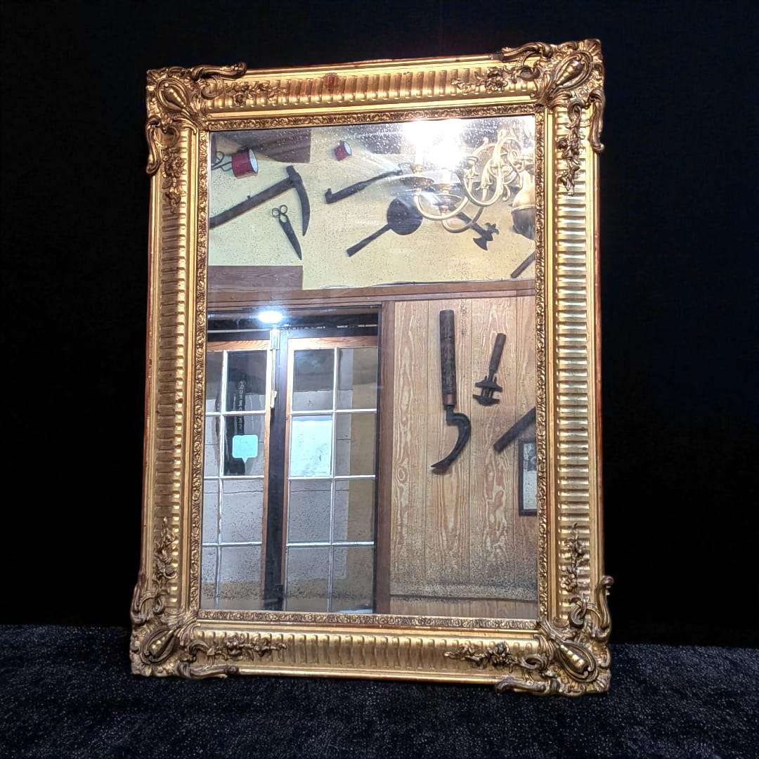 Espejo romántico antiguo con marco de pan de oro original colgado en pared blanca, reflejando interior elegante con decoración clásica.