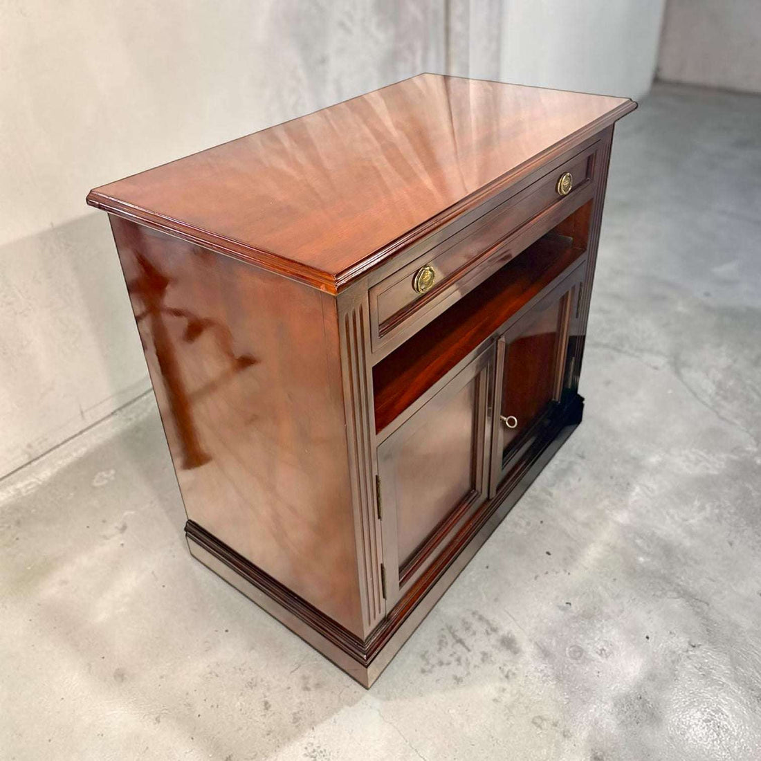 Medidas: 90 x 50 x H82,5 cm. Fabricada en caoba, esta Mesa Tv destaca por su acabado brillante en tono rojizo, su cajón superior con dos tiradores metálicos dorados, su hueco central abierto para aparatos o decoración y su alacena inferior con dos puertas y cerradura con llave.