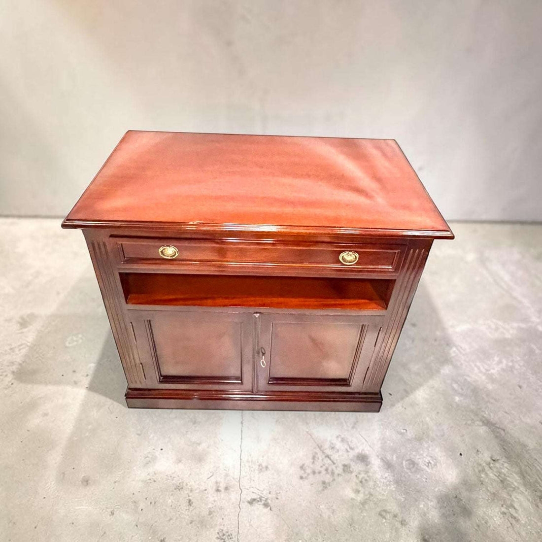 Medidas: 90 x 50 x H82,5 cm. Fabricada en caoba, esta Mesa Tv destaca por su acabado brillante en tono rojizo, su cajón superior con dos tiradores metálicos dorados, su hueco central abierto para aparatos o decoración y su alacena inferior con dos puertas y cerradura con llave.