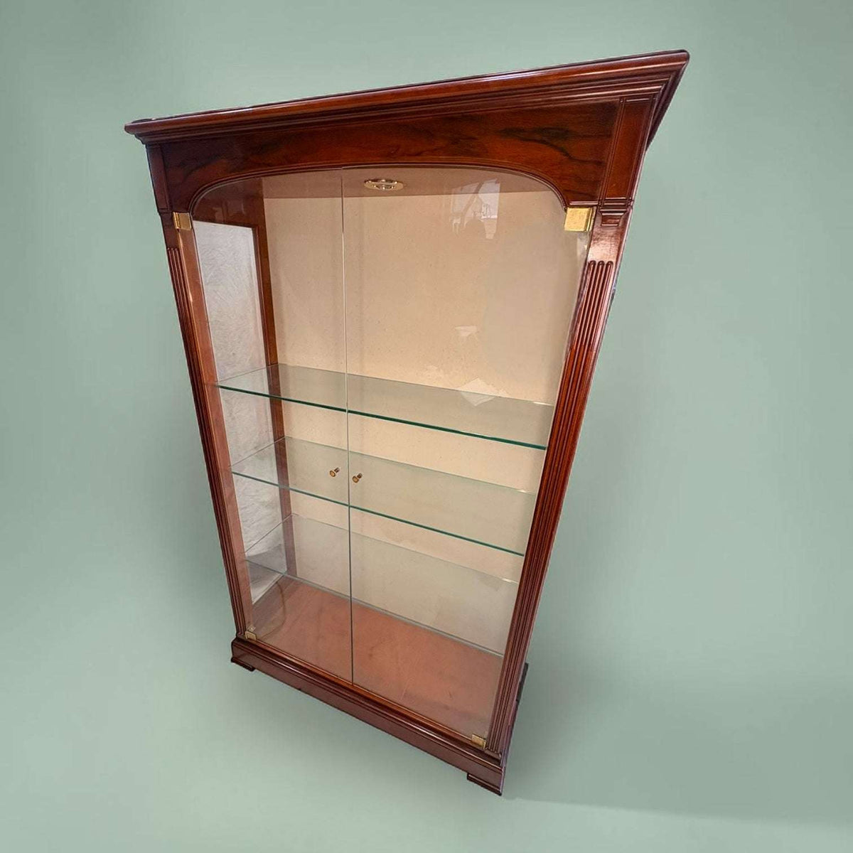 Esta Vitrina destaca por su diseño refinado, con una estructura alta y estilizada en madera de caoba de tono cálido y acabado brillante.