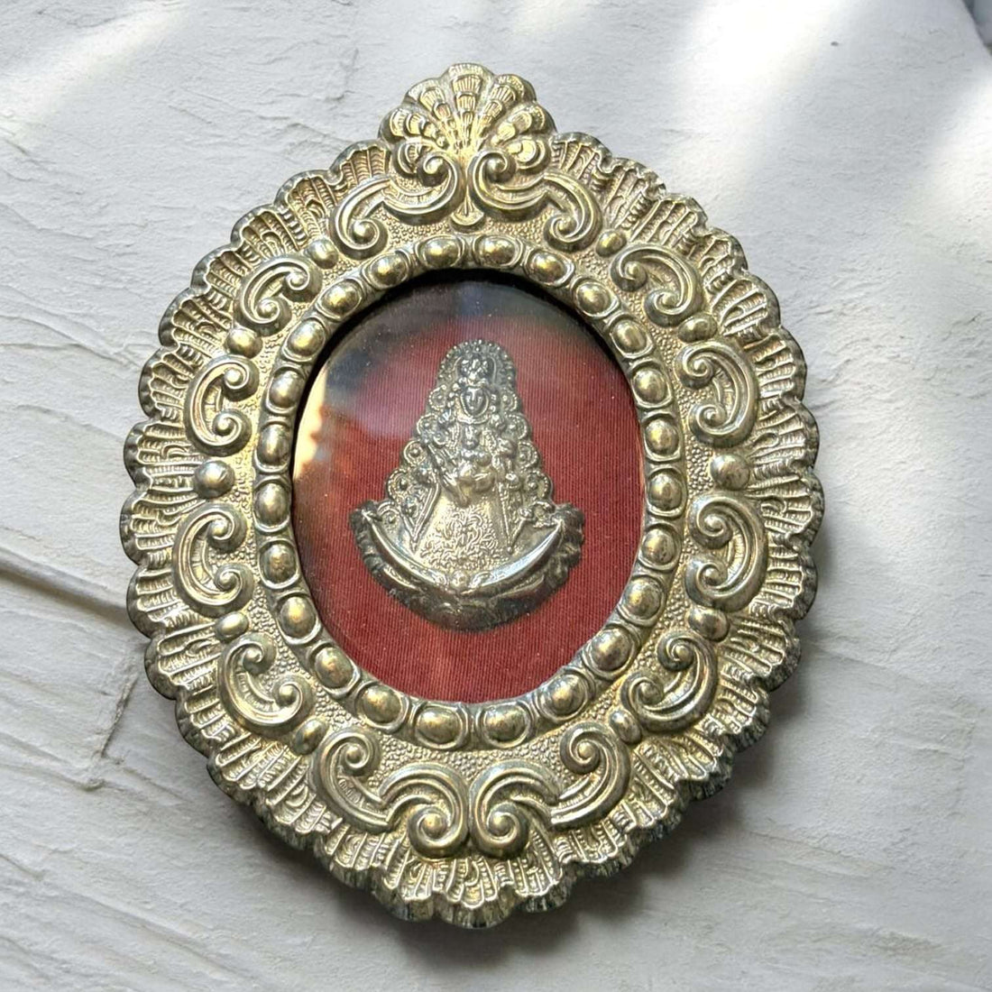 Este Relicario de metal plateado es una pieza devocional y decorativa de gran presencia, formada por un marco ovalado ornamentado y una imagen interior de la Virgen del Rocío sobre fondo textil rojo.