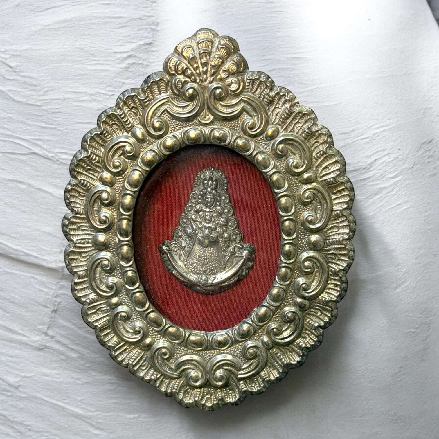 En el centro, la figura de la Virgen aparece en relieve metálico, protegida por cristal y realzada por el fondo rojo, que genera un contraste intenso y muy llamativo.