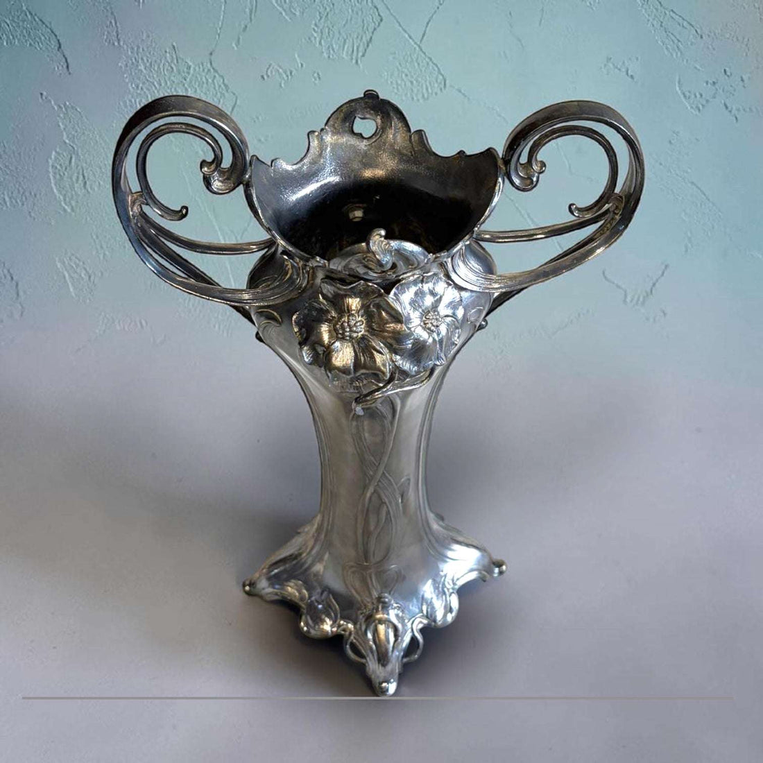 Este Florero estilo Art Nouveau, también conocido como Jugendstil, es una pieza decorativa de gran presencia visual, realizada en estaño con acabado plateado y diseño de inspiración vegetal. 