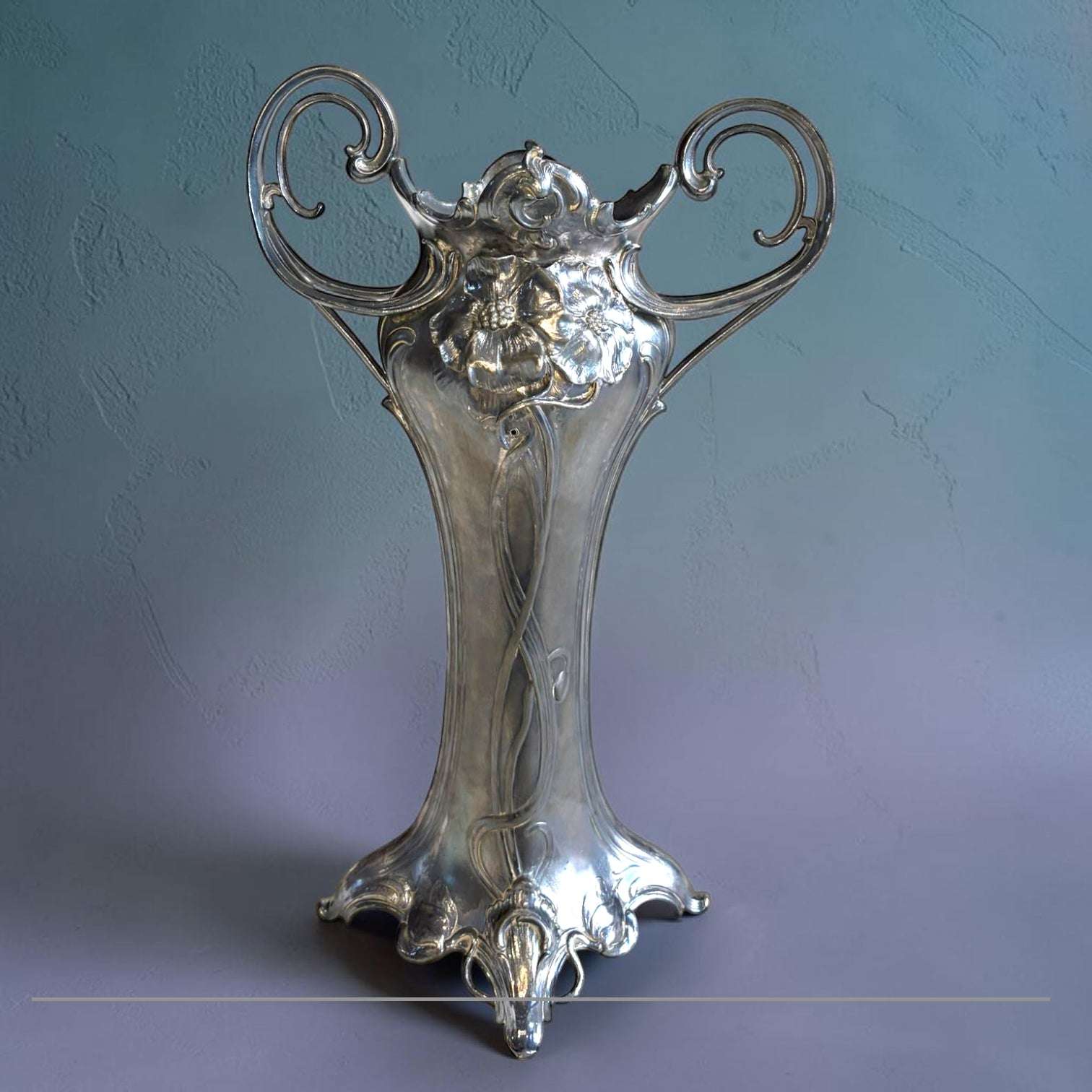 Este Florero estilo Art Nouveau, también conocido como Jugendstil, es una pieza decorativa de gran presencia visual, realizada en estaño con acabado plateado y diseño de inspiración vegetal. 