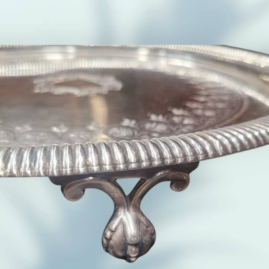Bandeja de metal plateado con ornamentos elegantes, diseño floral en relieve y escudo central, ideal para decoración vintage de interiores.