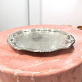 Bandeja ornamental de metal plateado con diseño vintage grabado, colocada sobre mesa de mármol rosa. Pieza decorativa para hogar elegante.