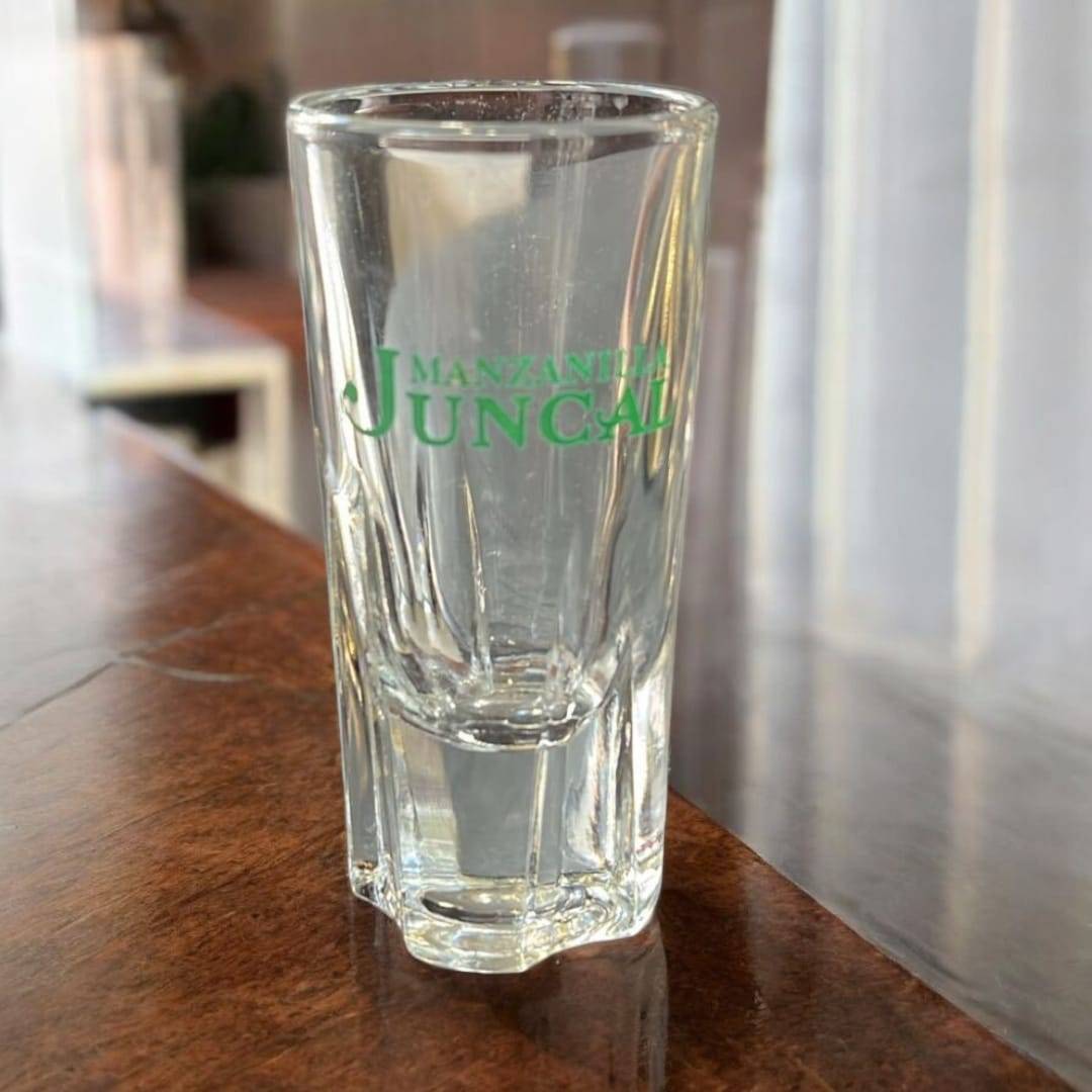 Cañero vintage de metal dorado con 12 vasos de cristal Juncal sobre mesa de madera. Ideal para decoración retro en hogar o bar.