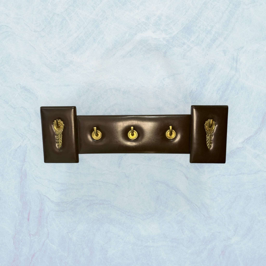 Perchero elegante de metal y cuero exclusivo y único con cuatro ganchos dorados y base marrón, ideal para decoración de interiores con estilo clásico.