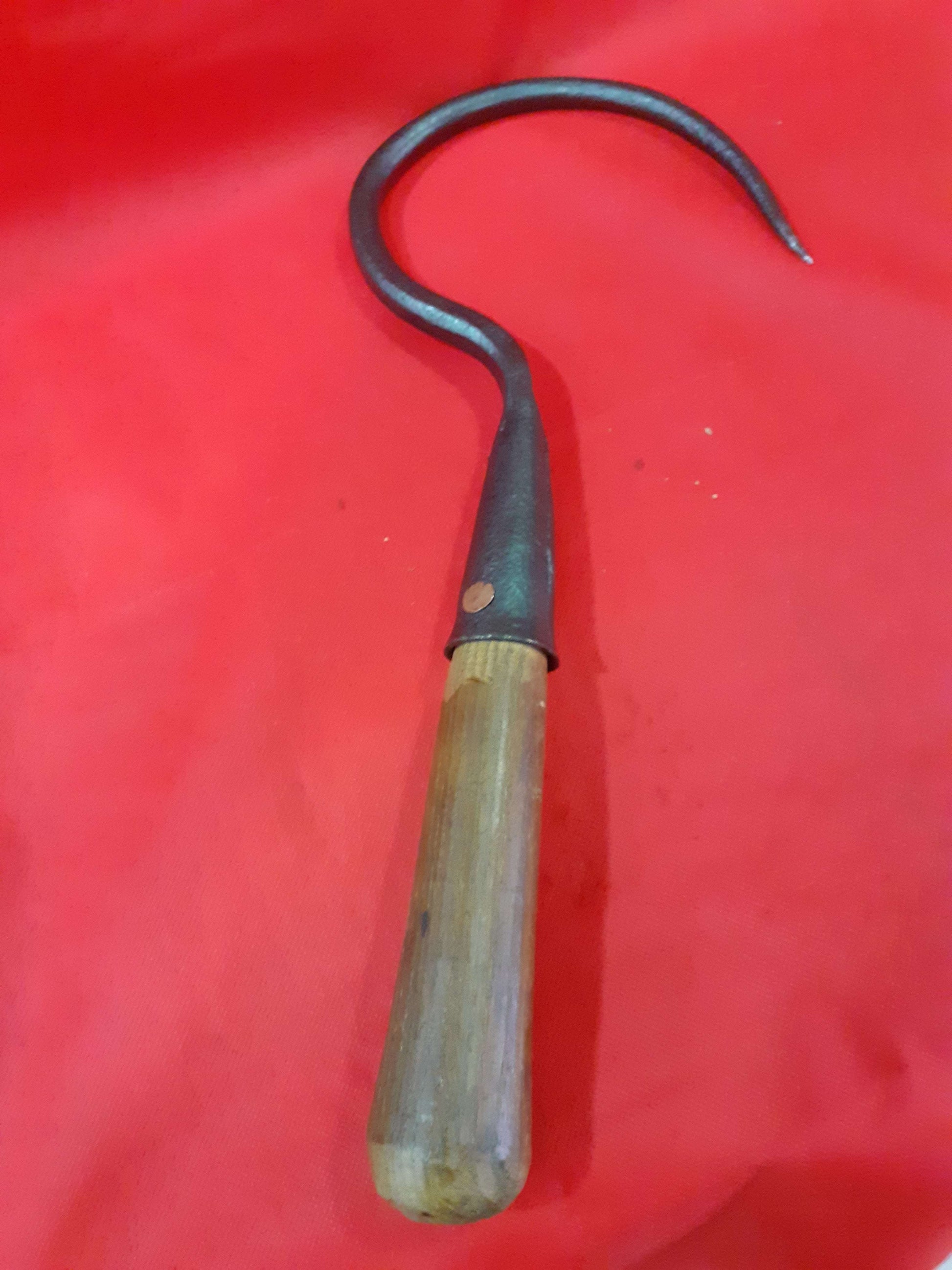 Garfio de hierro antiguo con mango de madera sobre fondo rojo, ideal para decoración rústica o coleccionistas. Disponible en Cositas Antiguas.