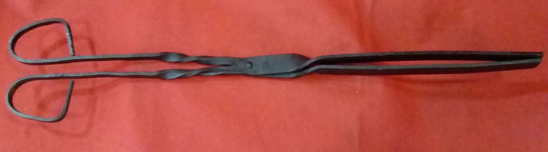 Pinza para brasero en hierro forjado sobre fondo rojo, ideal para decoración rústica y colecciones vintage de herramientas antiguas.