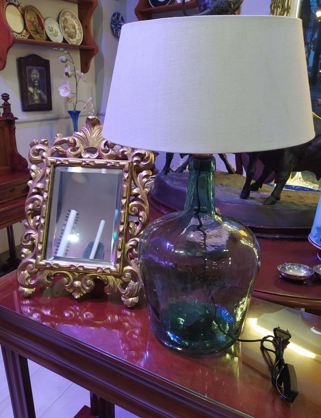 Lámpara de sobremesa de damajuana antigua de cristal verde con pantalla blanca, ideal para decoración vintage en interiores elegantes. Perfecta para iluminar espacios rústicos en el hogar.