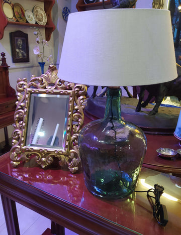 Lámpara de sobremesa de damajuana antigua de cristal verde con pantalla blanca, ideal para decoración vintage en interiores elegantes. Perfecta para iluminar espacios rústicos en el hogar.