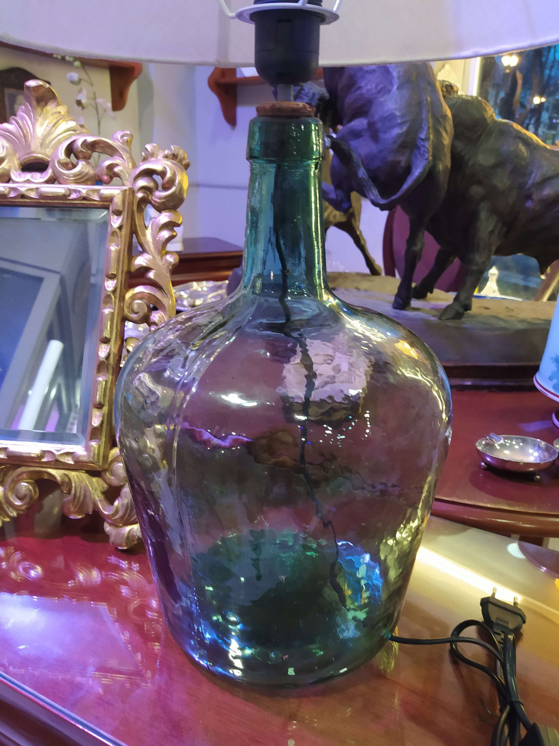 Lámpara de sobremesa de damajuana antigua de cristal verde con pantalla blanca, ideal para decoración vintage en interiores elegantes. Perfecta para iluminar espacios rústicos en el hogar.