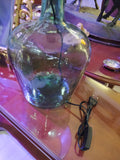 Lámpara de sobremesa de damajuana antigua de cristal verde con enchufe visible, ideal para decoración vintage en interiores elegantes. Perfecta para añadir un toque rústico y sofisticado en espacios del hogar.